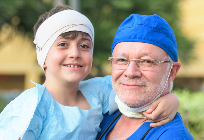 רופא מנתח מחזיק על ידיו ילד מחייך עם תחבושת על ראשו לאחר ניתוח. נוירוכירורגיה ילדים